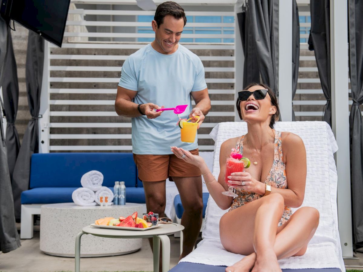 A man serves a colorful drink to a smiling woman lounging on a cabana bed, with a tray of snacks and towels on a side table.