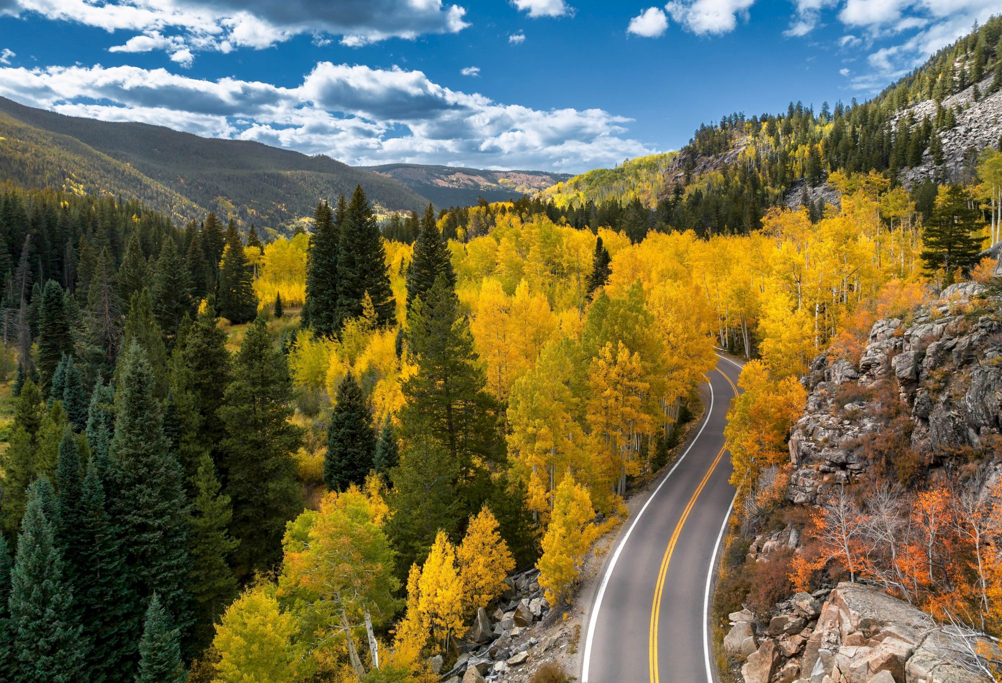 A winding road cuts through a vibrant autumn forest, with trees in shades of yellow and green under a partly cloudy sky.
