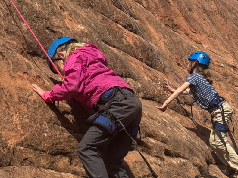 Two people rock climbing on a steep orange cliff, wearing helmets and harnesses, with ropes and climbing gear visible.