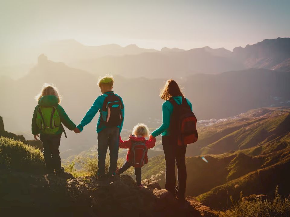 A family of four stands on a hilltop at sunrise, holding hands, overlooking rolling valleys and distant mountains on a hiking adventure.