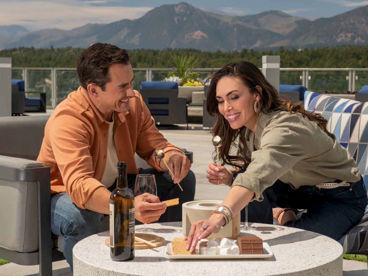 A couple enjoys a rooftop hangout with drinks and snacks on a sunny day, mountains in the background.