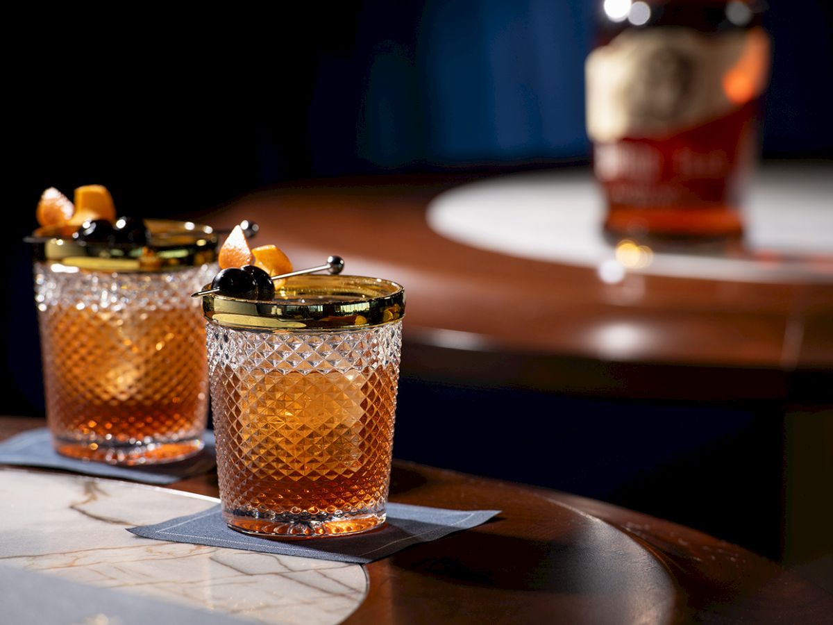 Two old-fashioned cocktails on a bar, garnished with citrus twists, on oak-topped glassware with a bottle in the blurred background.