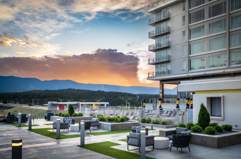 A modern rooftop lounge at sunset: comfy seating, glass railings, planters, and a view of distant mountains under a colorful sky.