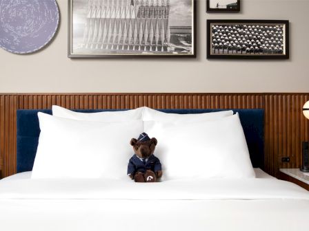 A cozy hotel bed with white sheets and pillows, a small teddy bear sitting upright in the center, framed photos on the wall above.