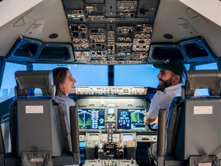 Two pilots sit in a cockpit, smiling and talking, surrounded by screens and controls in the aircraft cockpit.