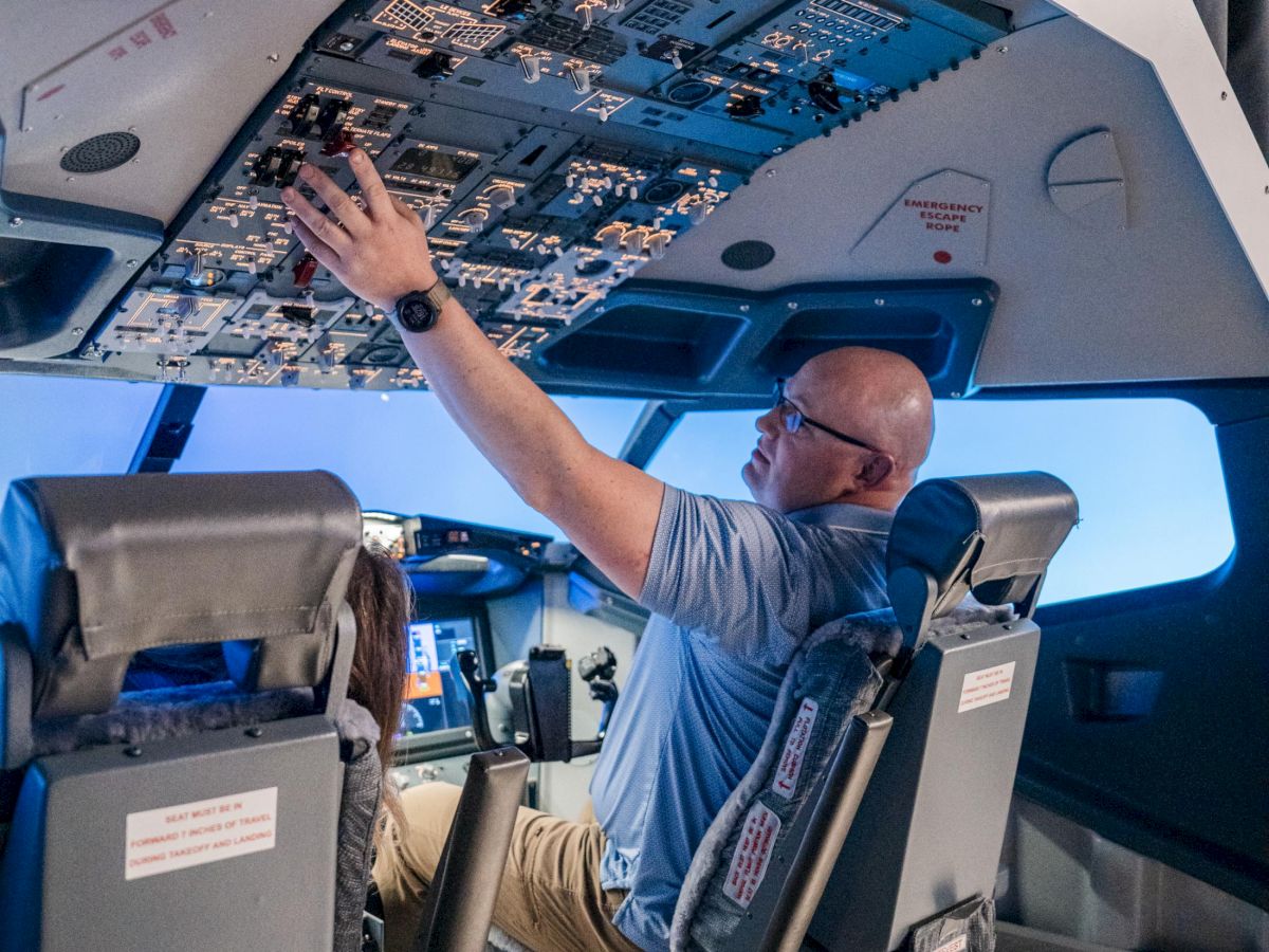 A pilot sits in the cockpit, reaching up to a ceiling panel with instruments and controls, inside a modern aircraft cabin.