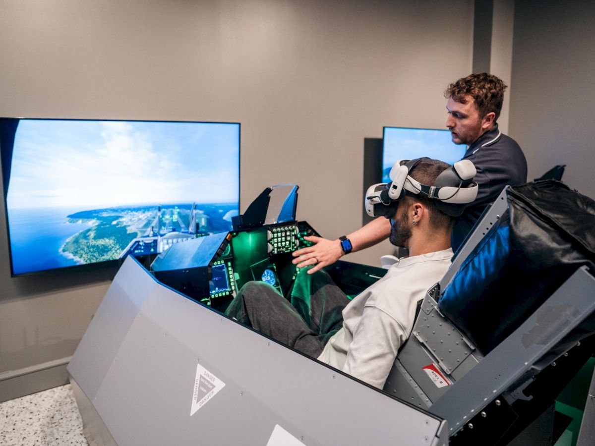 Two people in a cockpit-like simulator with large screens showing an ocean view; one is wearing a VR headset and controls, while a trainer stands beside.