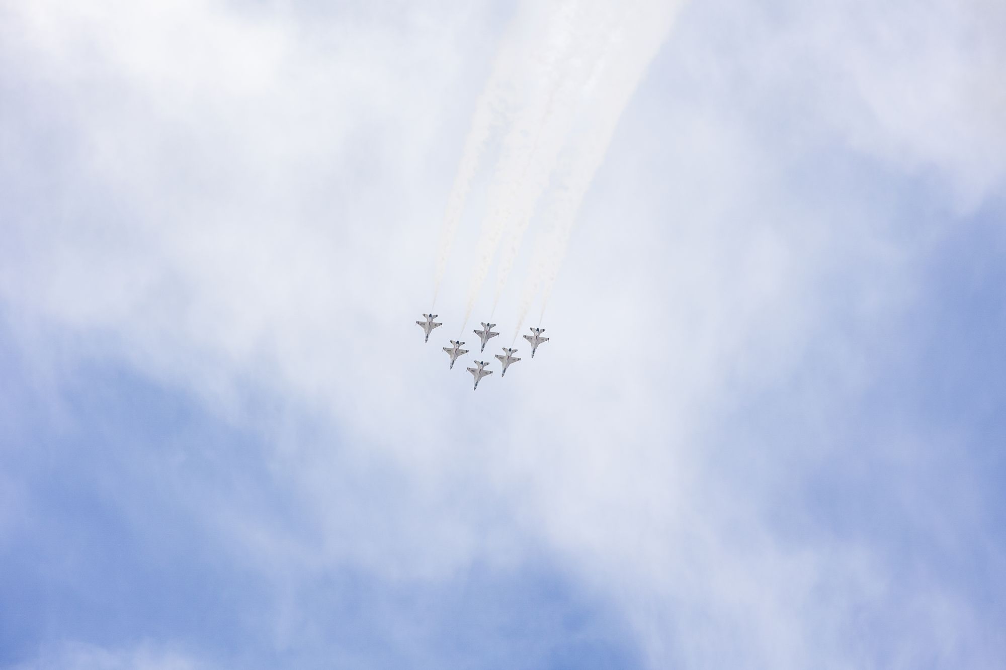 The image shows a formation of jets flying in the sky, leaving vapor trails against a backdrop of clouds.