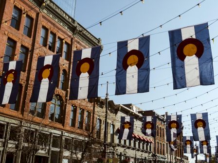 Blue banners with a white vertical stripe and a gold circle hang across a street, strung with lights above brick storefronts, festive urban decor.