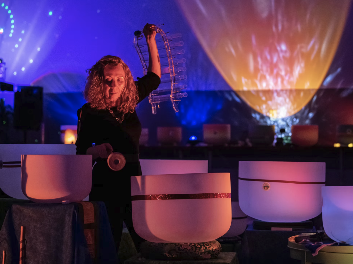 A woman performs in front of glowing stage lights, beside harp-like instruments and a microphone during a live music show.