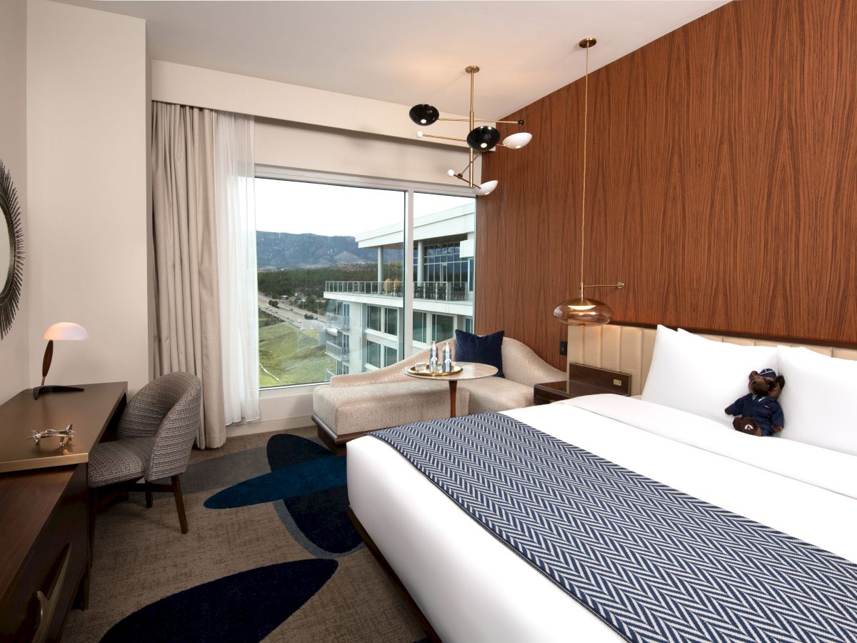 The image shows a modern hotel room with a large bed, desk, chair, a window with a view, and a teddy bear on the bed.