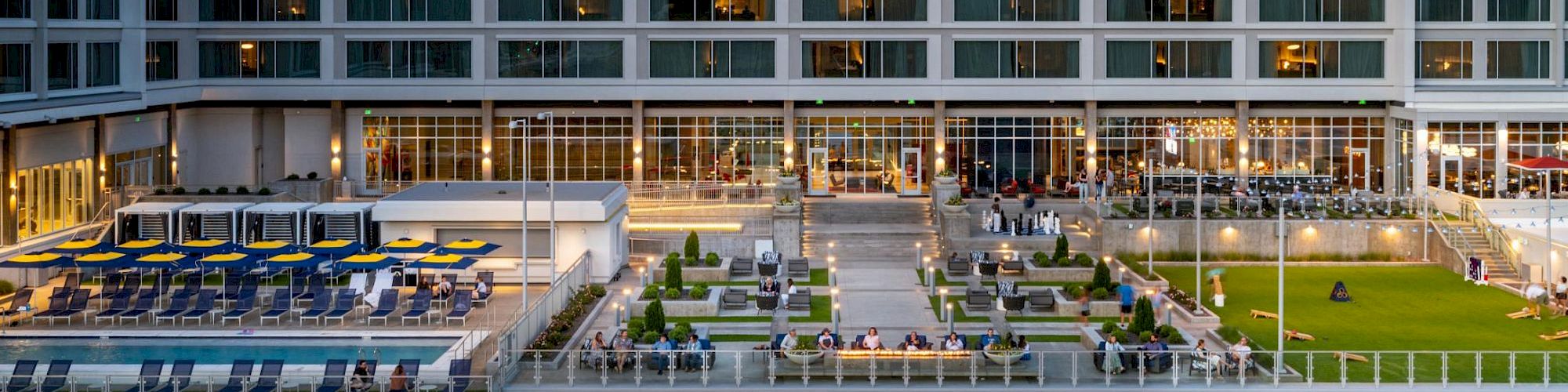 A modern hotel courtyard at dusk with a pool terrace, striped umbrellas, lit windows, and lounge seating outside.