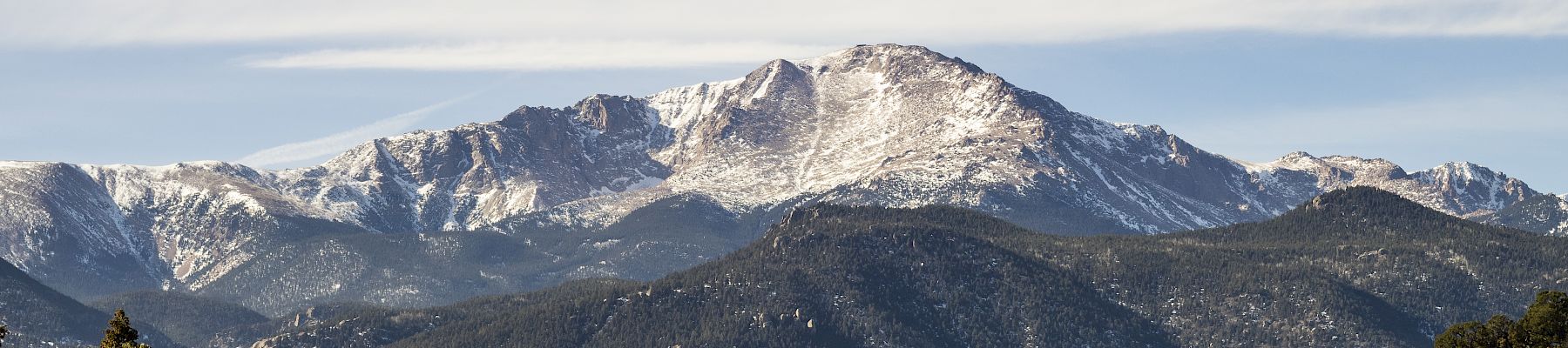 A snow-capped mountain range under a blue sky with scattered clouds, surrounded by forested hills and valleys.