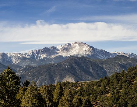 A snow-capped mountain range under a blue sky with scattered clouds, surrounded by forested hills and valleys.