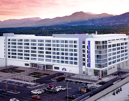 A modern, multi-story building with a parking lot, set against mountains and a colorful sky in the background.