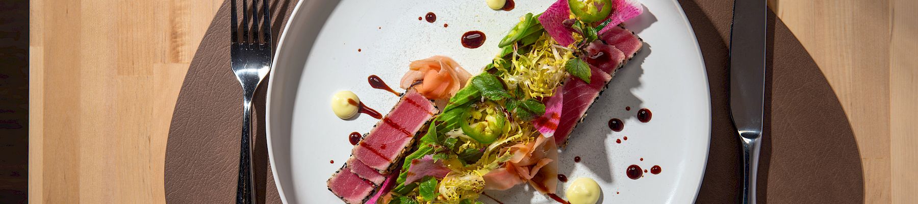 A plated dish with sliced meat, sauce drops, garnishments, with utensils and a glass on a wooden table.