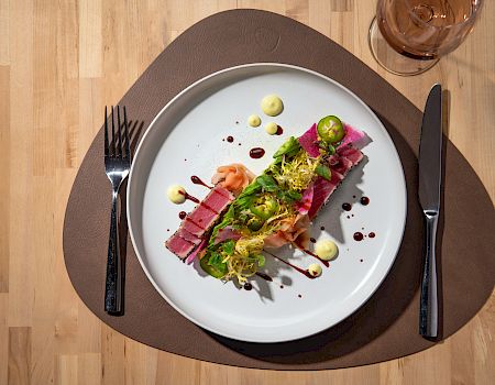 A plated dish with sliced meat, sauce drops, garnishments, with utensils and a glass on a wooden table.