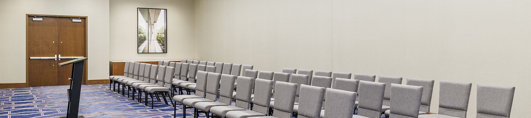 An empty conference room with rows of gray chairs, a podium, patterned blue carpet, and doors at the back wall.