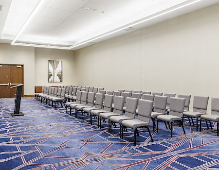 An empty conference room with rows of gray chairs, a podium, patterned blue carpet, and doors at the back wall.