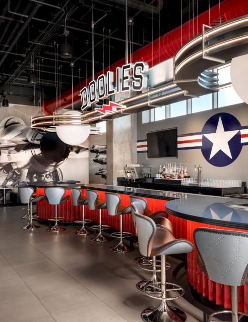 A retro-style diner bar with red curved counter, modern gray-and-red stools, star-spangled decor, airplanes mural, and a large sign reading &ldquo;Doubles&rdquo; overhead.