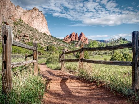 A dirt trail winds through a wooden fence in a sunny valley, with rugged red rock formations and blue sky in the background.
