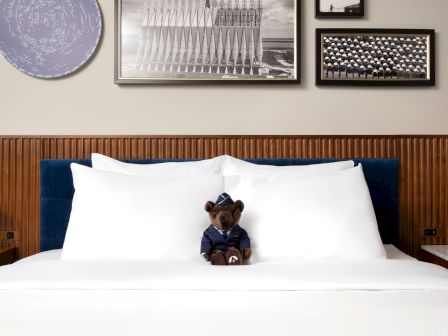 A teddy bear in uniform sits on a neatly made bed with white pillows, surrounded by framed photos on the wall.