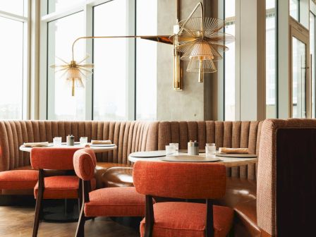 The image shows a stylish restaurant interior with round tables, orange chairs, and modern lighting fixtures near large windows.