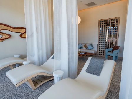 The image shows a relaxing spa room with lounge chairs, white curtains, wall art, and a seating area with cushions and soft lighting.