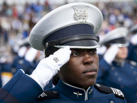A person in military uniform performs a salute, with others similarly dressed in the background.