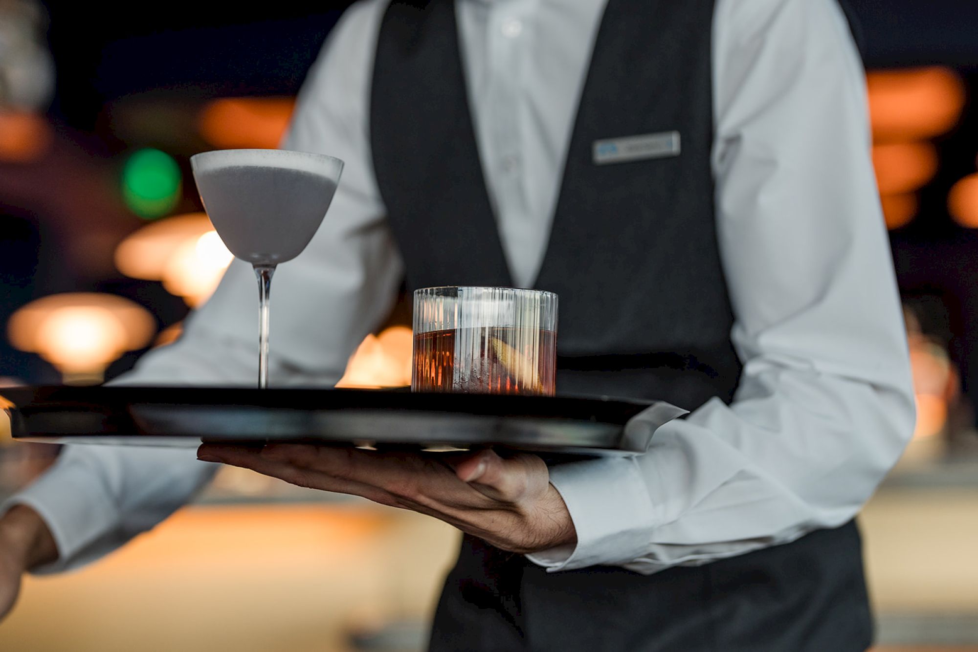 A waiter in a vest carries a tray with a martini glass and a tumbler with a drink and lemon slice in a dimly lit setting.