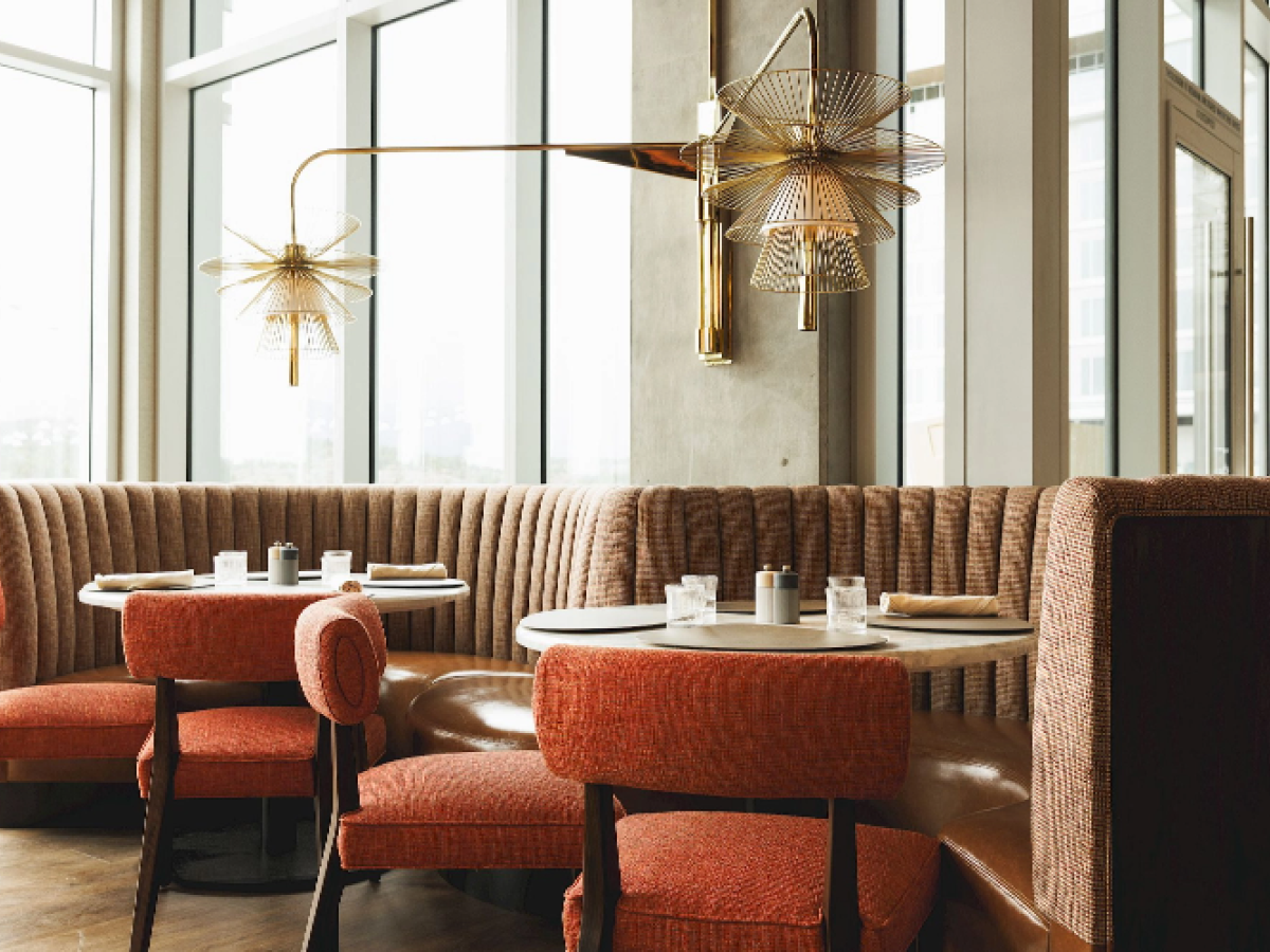 A cozy restaurant interior with round tables, upholstered chairs, booth seating, and decorative pendant lighting by large windows.