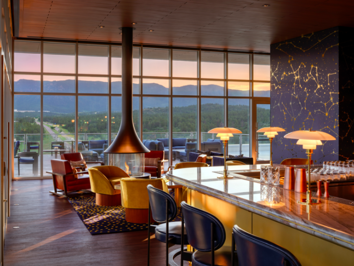 A modern lounge with a bar, cozy seating, large windows showing mountains, and a hanging fireplace, exuding a warm and stylish ambiance.