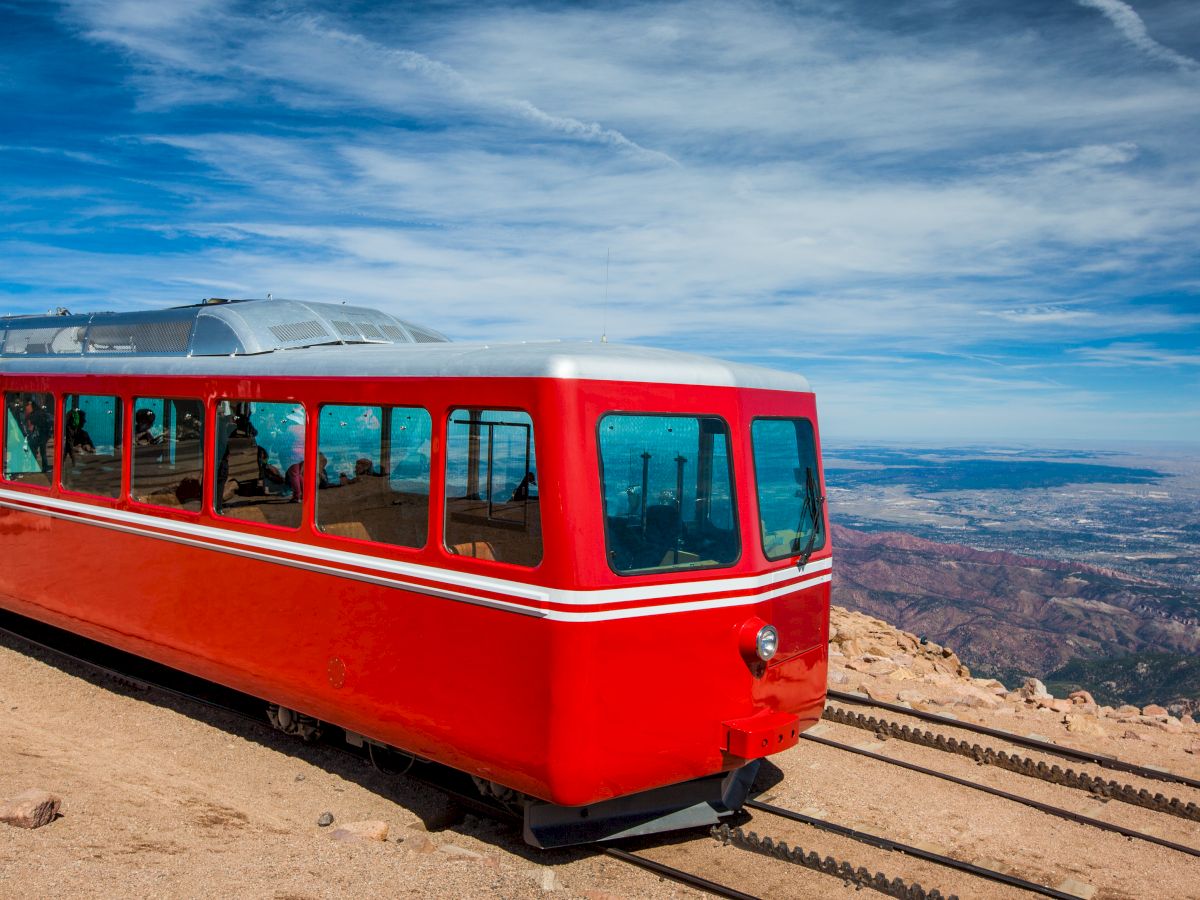 A red cog railway train is on a mountain track with a scenic view of the landscape and blue sky in the background.