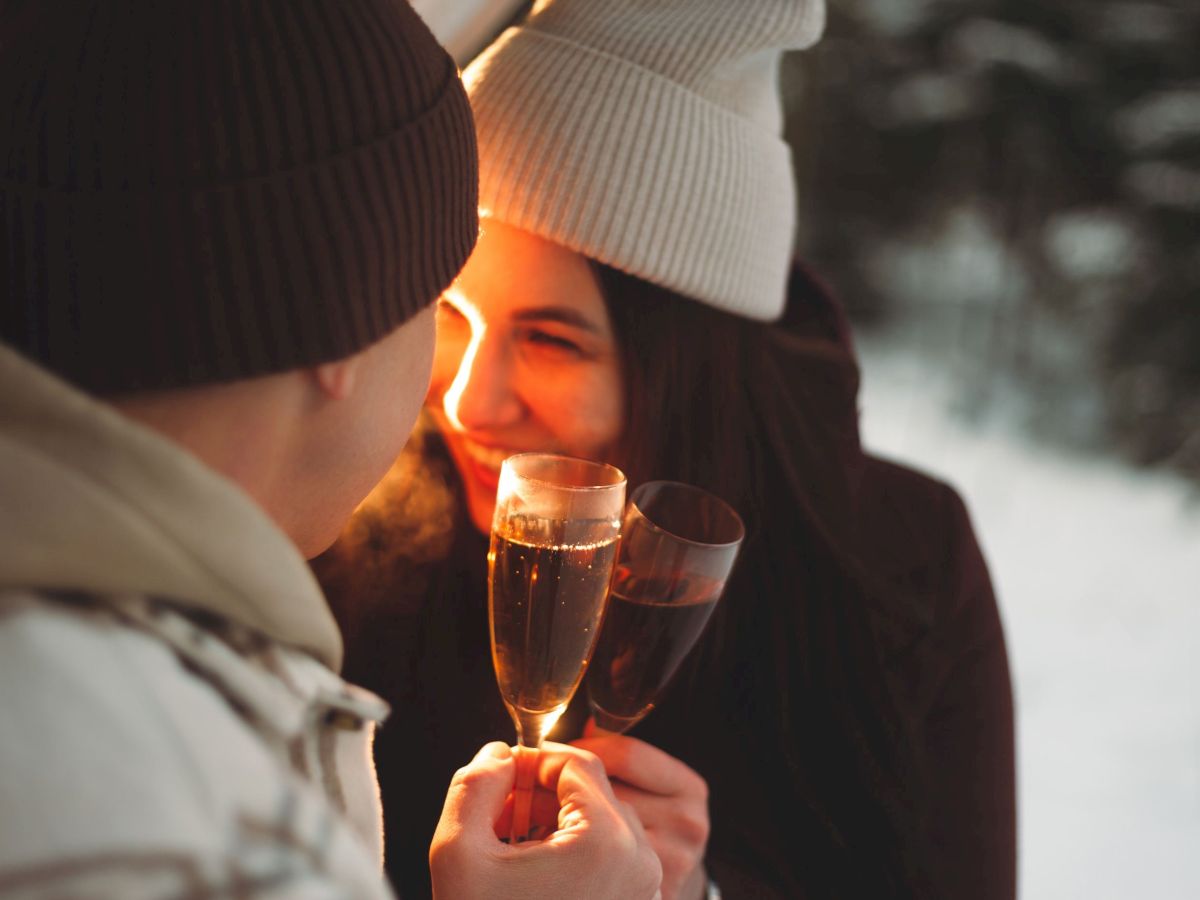 Two people in winter attire smile warmly, clinking glasses of champagne outdoors, surrounded by a snowy backdrop.