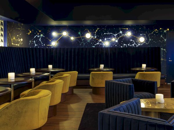 A stylish lounge with dark velvet seating, round tables, yellow chairs, and starry wall decorations, lit by soft ambient lights.