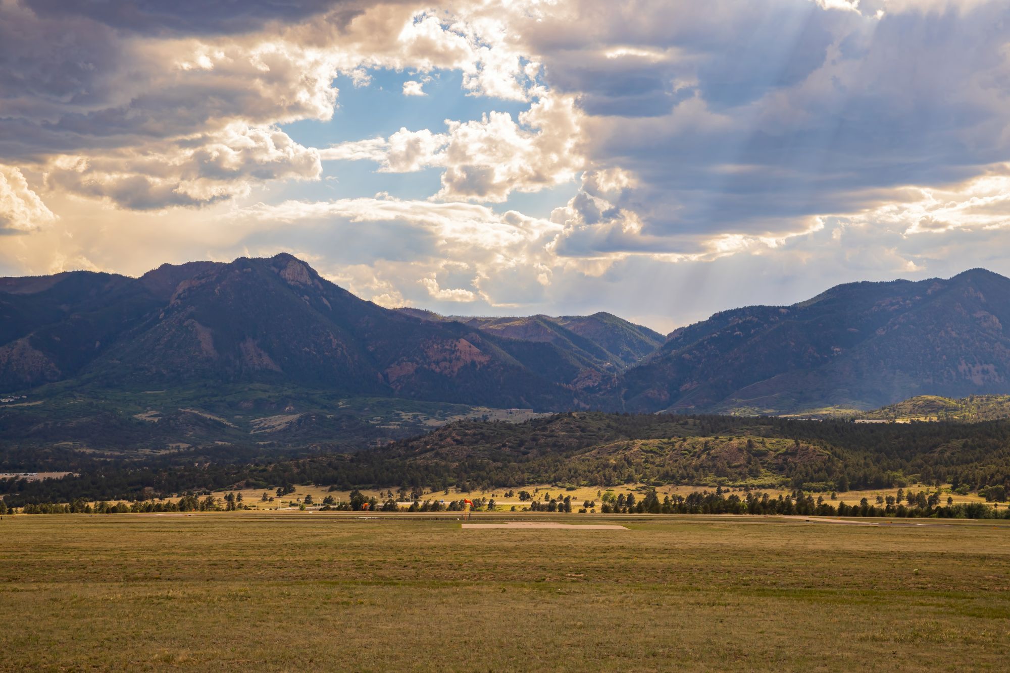 A vast field stretches towards distant mountains under a partly cloudy sky, with sunlight breaking through in patches.