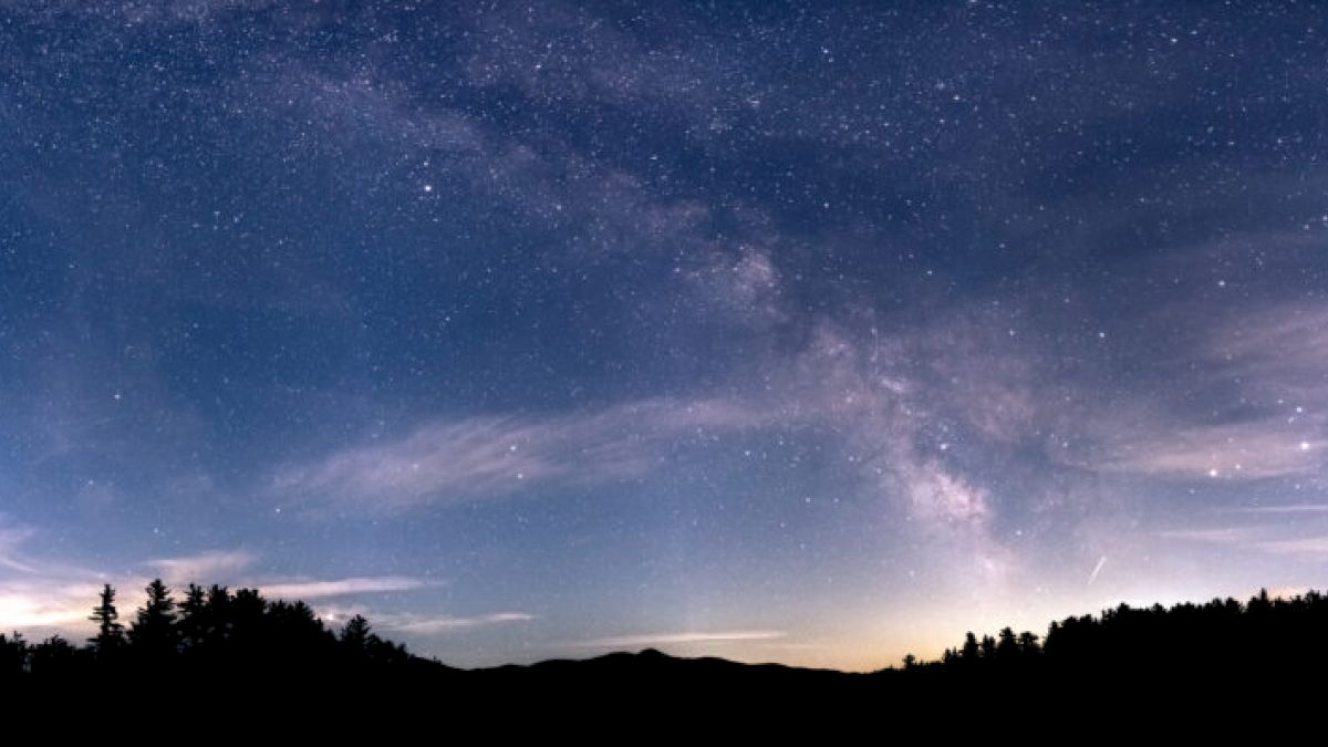 A starry night sky with the Milky Way visible, silhouetted trees in the foreground, and faint clouds adding texture to the celestial scene.