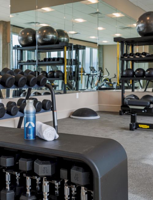 A gym with dumbbells, exercise equipment, mirrors, a water bottle, and a towel on a rack, creating a well-equipped workout space.