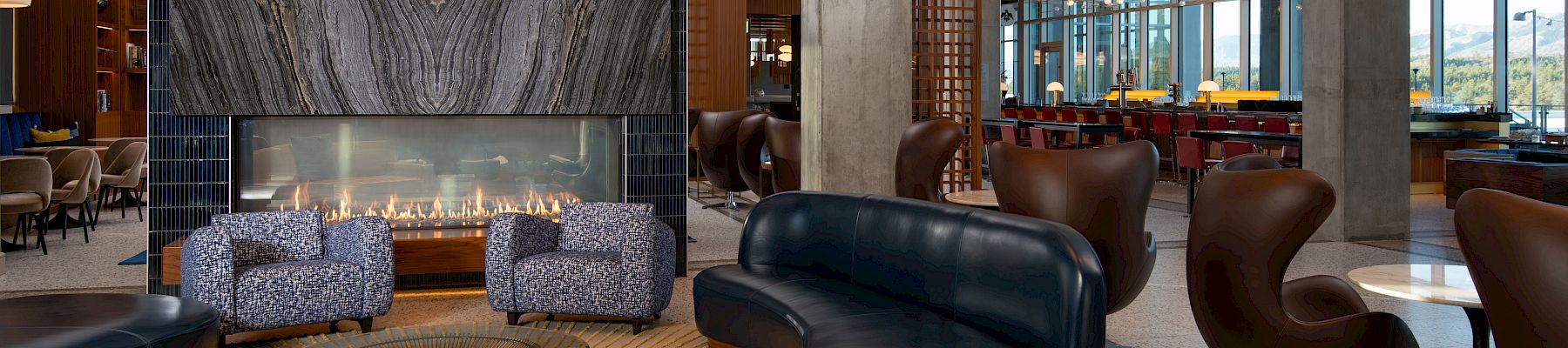 A modern lounge area with patterned chairs, a sculptural sofa, a large stone fireplace, and open spaces featuring natural light and chic decor.