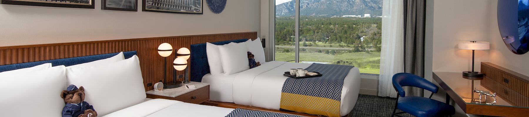 The image shows a hotel room with two beds, a mountain view, modern decor, and a teddy bear on one bed.