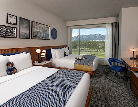 The image shows a hotel room with two beds, a mountain view, modern decor, and a teddy bear on one bed.