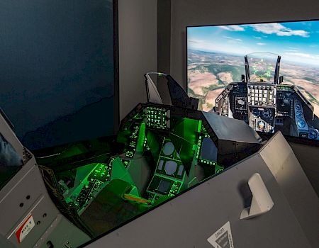 A flight simulator cockpit with illuminated controls is shown, featuring a large screen displaying a realistic aerial landscape view.