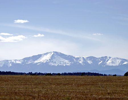 The image shows a snow-capped mountain range under a clear blue sky, with a flat grassy field in the foreground.