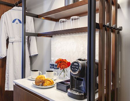 A cozy setup with a coffee machine, cups, croissants, flowers, glasses, and a hanging robe on a shelf.