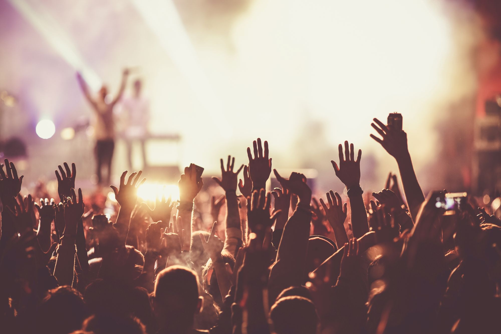 A crowd at a concert with hands raised in the air, bright stage lights in the background.