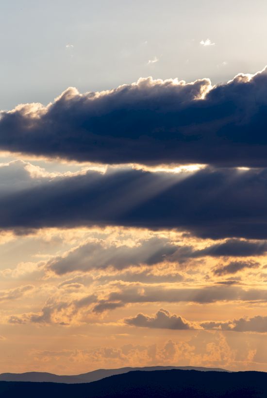 A scenic view of a sunset with clouds and sun rays illuminating the sky above a distant mountain horizon.