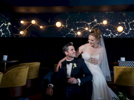 A bride and groom sit in a stylish room with constellation wall art and warm lighting, sharing a joyful moment together.