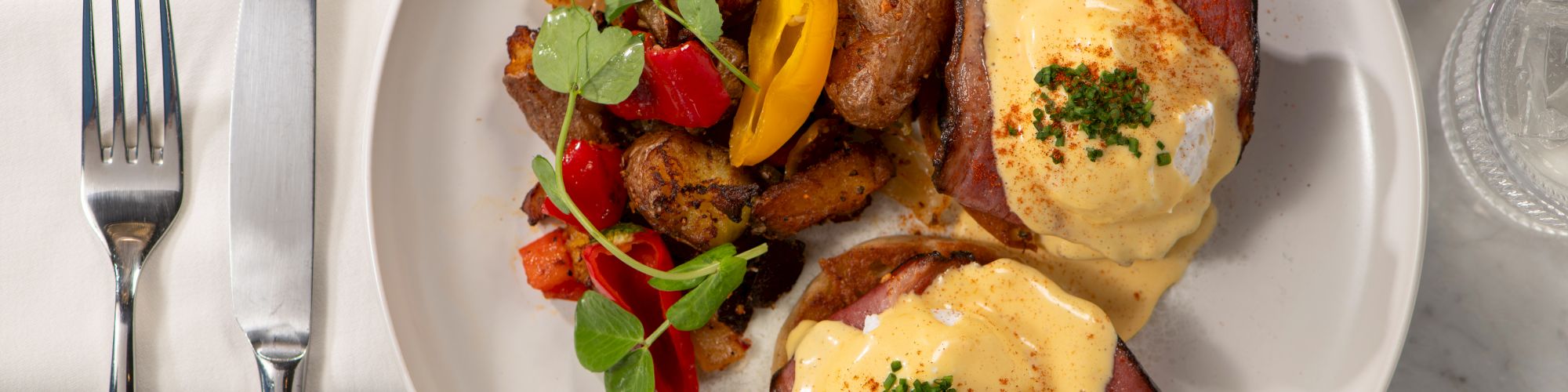 The image shows eggs Benedict with hollandaise sauce, home fries, and greens on a white plate, accompanied by cutlery and a napkin.