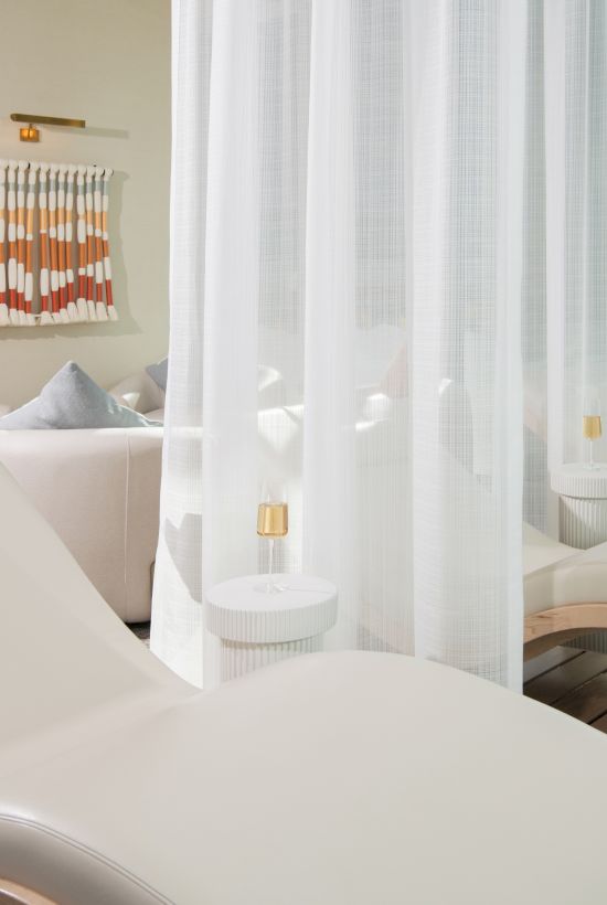 The image shows a spa-like setting with white cushioned lounge chairs, a curtain, and modern decor, including a colorful wall hanging.