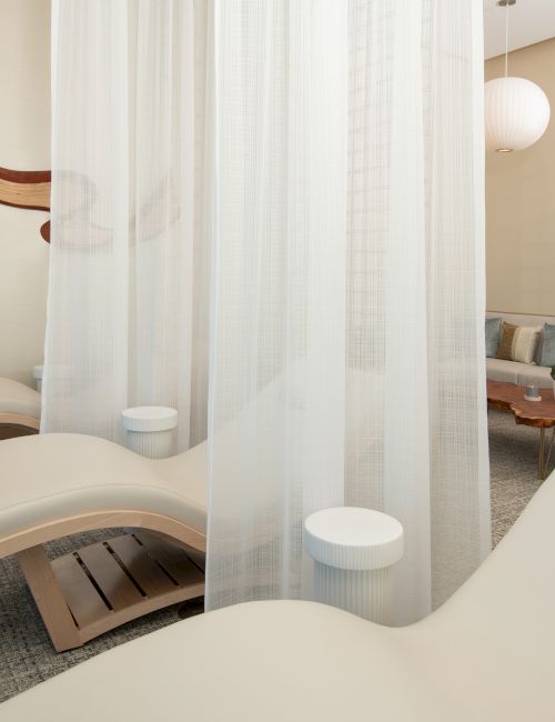The image shows a serene spa room with comfortable lounge chairs, sheer curtains, and soft lighting for relaxation and tranquility.
